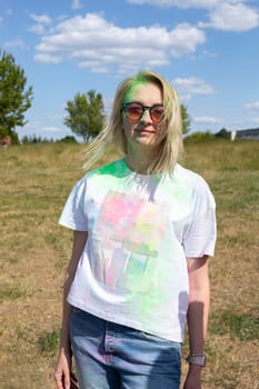 Smiling Beautiful White Blond Woman With Colorful Dye, Powder On Face, Cloth On Holi Colors Festival In Park, Sunny Day. Playful Cultural Event With Throwing Bright Neon Powder. Vertical Plane.