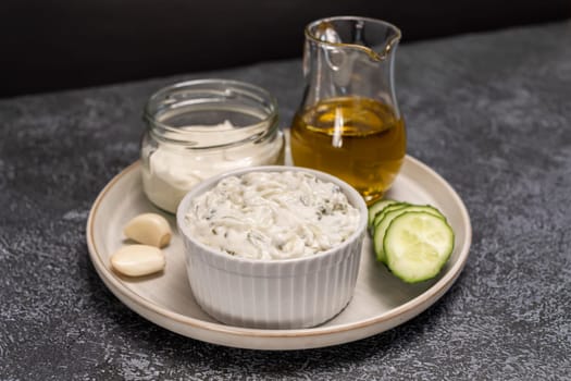 Traditional greek yogurt sauce Tzatziki in white bowl, made of chopped cucumbers, sour cream yogurt,olive oil,garlic,herbs with ingredients on on dark gray concrete table. Horizontal plane.