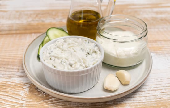 Traditional greek yogurt sauce Tzatziki in white bowl, made of chopped cucumbers, sour cream yogurt,olive oil,garlic,herbs with ingredients on wooden table. Rustic style. Close up, horizontal plane.
