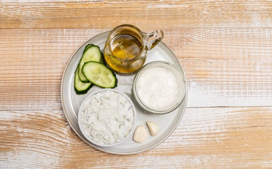 Traditional greek yogurt sauce Tzatziki in white bowl, made of chopped cucumbers, sour cream yogurt,olive oil,garlic,herbs with ingredients on wooden table. Rustic style. Horizontal plane, top view