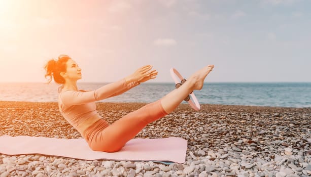 Middle aged well looking woman with black hair doing Pilates with the ring on the yoga mat near the sea on the pebble beach. Female fitness yoga concept. Healthy lifestyle, harmony and meditation.