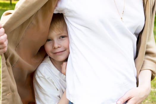 Cute Smiling White Boy Looks Out Mother's Jacket, Hiding Behind Her Back In Park. Mom's Support And Care. Summer Time. Parenthood, Family Leisure Time. Children's Day. Horizontal Plane.