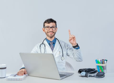 Smiling doctor with laptop with finger up in idea gesture. Doctor sitting with laptop pointing up isolated. Young doctor sitting with laptop with an idea gesture.