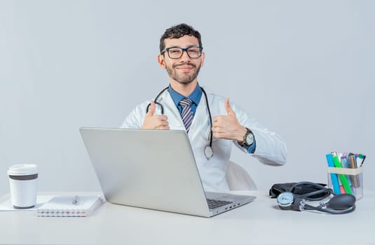 Smiling doctor sitting with laptop giving thumbs up. Male doctor with laptop showing thumbs up in ok gesture.