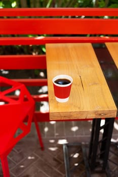 A wooden table on which there is a red plastic cup with coffee. Summer beautiful terrace in greenery