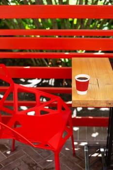 A wooden table on which there is a red plastic cup with coffee. Summer beautiful terrace in greenery. Cozy place
