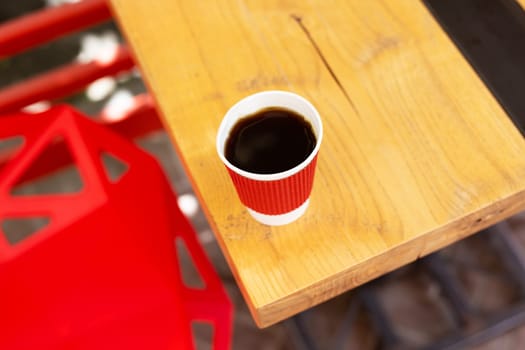 A wooden table on which there is a red plastic cup with coffee. Summer beautiful terrace in greenery. Cozy place