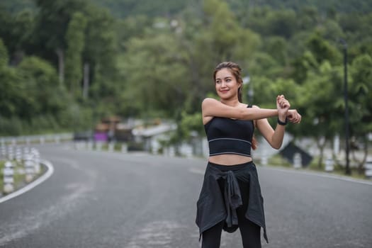 A portrait of a sporty and happy young Asian woman in sportswear is stretching her arms, warming up her body before running outdoors. Outdoor activity concept..