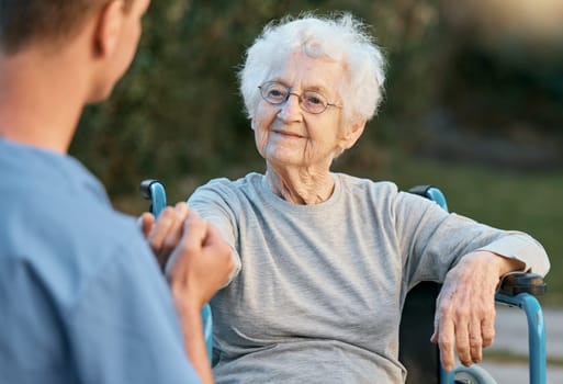 Senior woman, wheelchair and caregiver holding hands with patient for empathy, support and care outdoor for communication and air. Man nurse and old woman talking rehabilitation at nursing home.