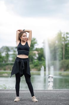 A portrait of a sporty and happy young Asian woman in sportswear is stretching her arms, warming up her body before running outdoors. Outdoor activity concept..