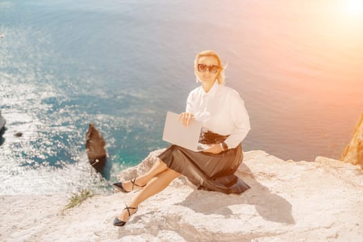 Business woman on nature in white shirt and black skirt. She works with an iPad in the open air with a beautiful view of the sea. The concept of remote work