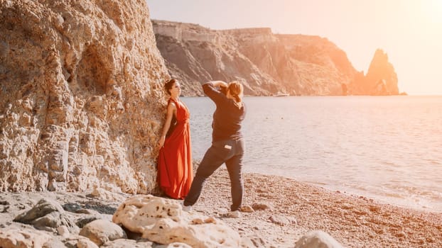 Side view a Young beautiful sensual woman in a red long dress posing on a rock high above the sea during sunrise. Girl on the nature on blue sky background. Fashion photo.