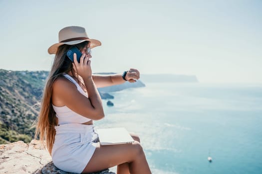 Successful business woman in yellow hat working on laptop by the sea. Pretty lady typing on computer at summer day outdoors. Freelance, travel and holidays concept.