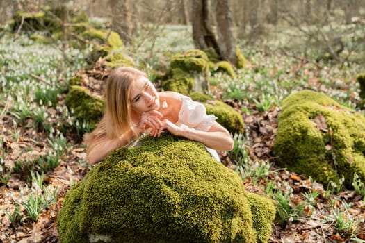 Snowdrops galanthus blond. A girl in a white dress lay down on a stone in the moss in a meadow with snowdrops in a spring forest.