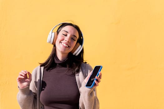 woman dancing happy listening to music from phone in headphones with a yellow wall in the background, concept of rhythm and positive people, copy space for text