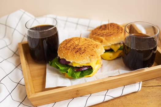 Big and tasty burgers lie on a wooden tray with a glass of cola. Wooden table and white checkered napkin. Close-up