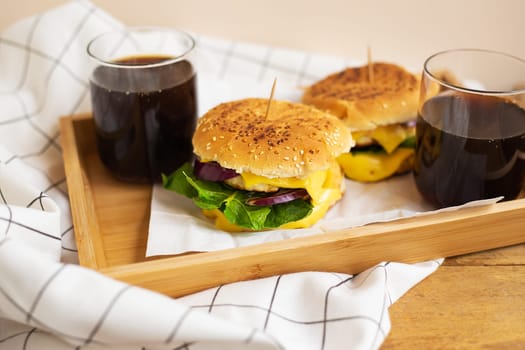 Big and tasty burgers lie on a wooden tray with a glass of cola. Wooden table and white checkered napkin. Close-up