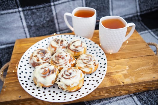 Two cups of black tea stand on a wooden tray on the sofa with a black and white checkered plaid. Fresh and fragrant cinnamon rolls close up lie on a plate with polka dots, beautiful morning.