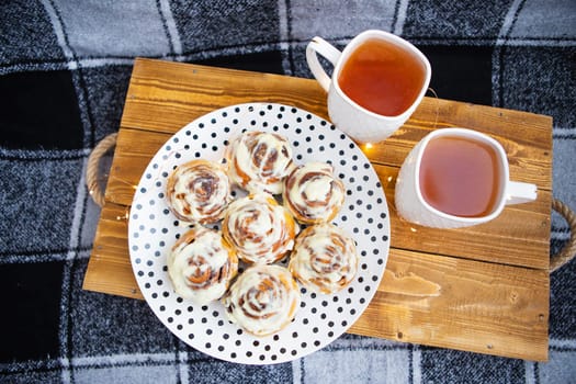 Two cups of black tea stand on a wooden tray on the sofa with a black and white checkered plaid. Fresh and fragrant cinnamon rolls close-up lie on a plate with polka dots. The garland is blinking.