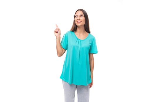 young brunette european lady with straight hair dressed in a blue t-shirt demonstrates with a hand gesture against a wall with advertising space.