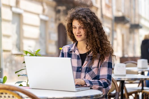 Young woman working online distant job with laptop in city sunshine street cafe or restaurant browsing website chatting outdoors during break. Girl tourist looking computer screen. Town lifestyles