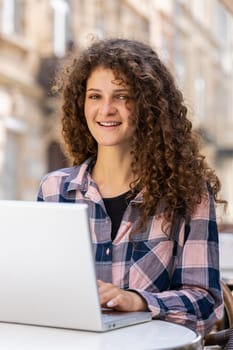 Young woman working online distant job with laptop in city sunshine street cafe or restaurant browsing website chatting outdoors during break. Girl tourist looking at camera. Town lifestyles. Vertical