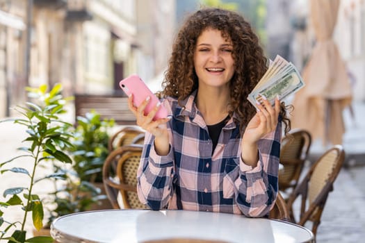 Happy rich woman counting money dollar cash, use smartphone calculator app, plans to order gifts, food delivery online, booking hotel room outdoors. Girl tourist sitting at table in city cafeteria