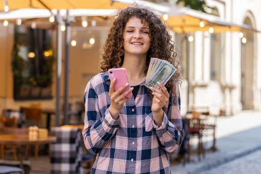Happy rich young woman counting money dollar cash, use smartphone calculator app, plans to order gifts and food delivery online, booking hotel room. Pretty girl tourist in city street. Town outdoors