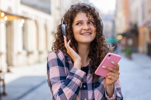 Happy relaxed overjoyed pretty young woman in wireless headphones choosing, listening favorite energetic disco rock n roll music dancing outdoors. Girl walking in urban city sunshine street background