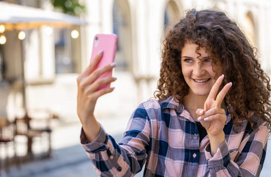 Pretty young woman blogger taking selfie on smartphone, communicating video call online with subscribers, recording stories for social media vlog outdoors. Girl walking in urban sunshine city street