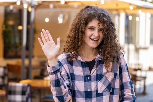 Hello. Young pretty woman smiling friendly at camera, waving hands gesturing hi, greeting, goodbye, welcoming with hospitable expression outdoors. Happy girl walking in urban sunshine city town street