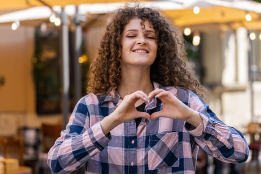 I love you. Young woman makes symbol of love, showing heart sign to camera, express romantic feelings, express sincere positive feelings. Charity, gratitude, donation. Outdoors in urban city street