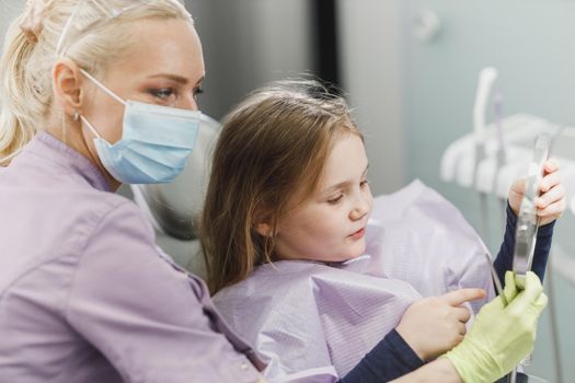 A female dentist and cute little girl's holding a mirror and looking her teeth after dental examination at dentist's office.