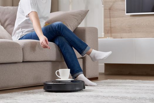 Robotic vacuum cleaner cleaning room while woman resting on sofa