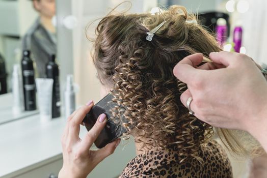 Hairdresser working with beautiful woman brown hair in hairdressing salon. Close up view of hand, curling iron and the appliance