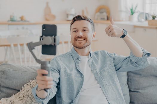 Young man in video call expressing his satisfaction, holding gimbal with phone attached, making thumbs up gesture and showing it to camera while sitting on couch, blurred background