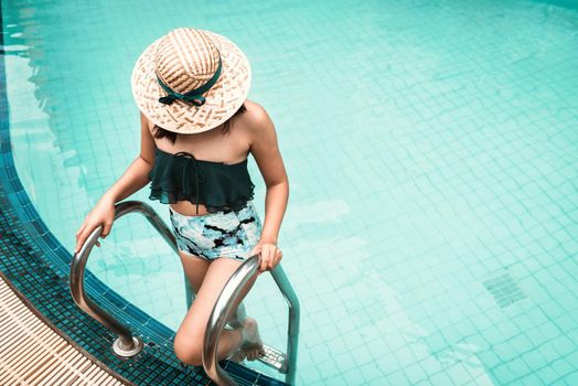 Sexy Woman in Swimsuit is Relaxing in Swimming Pool, Beautiful Woman Wearing Straw Hat and Relax in Poolside While Holding Pool Pedal Ladder at Resort Hotel. Summer Vacation and Relaxation Lifestyles