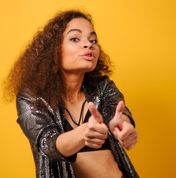 Thumbs up happy Afro American girl with beautiful hairstyle posing smile looking side away with hand on hips wearing shiny black jacket and black top on yellow background.