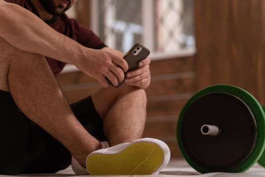 Young man texting sitting on a floor with smartphone, black and green tone fitness barbell, equipment for weight training concept. Healthy lifestyle concept. No face body shot. 