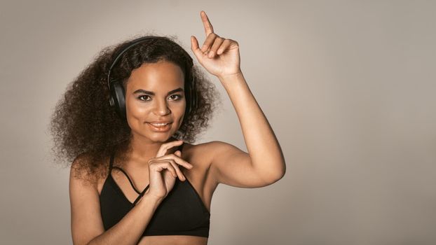 Rhythm dancing listening musing in headphones young African-American girl wearing black bare shoulders top and make salsa moves with hands in the air, isolated on grey background, emotionally move.