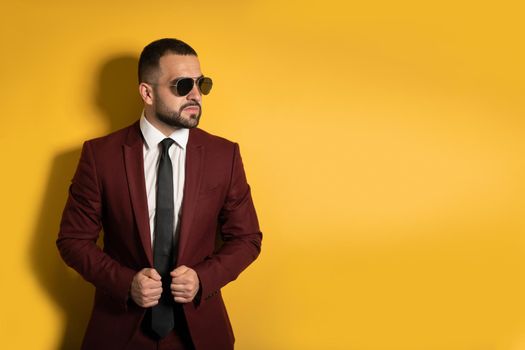 Young man in burgundy suit looking seriously sideways wearing sunglasses with hands holding a jacket isolated on yellow background. Men fashion concept. 