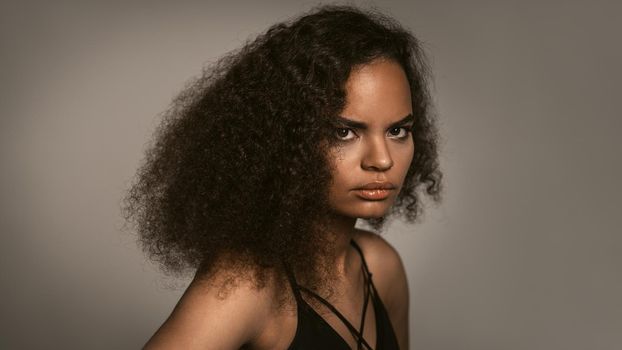 Passionate look of an African American young woman standing in black bare shoulder top isolated on grey background. Human emotions, facial expression concept.