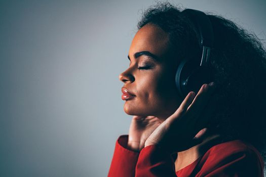 Joy of a music young African-American girl listening her favourite track in headphones wearing red jacket black top under isolated on grey background, emotionally move, have fun. Cold toned image. 