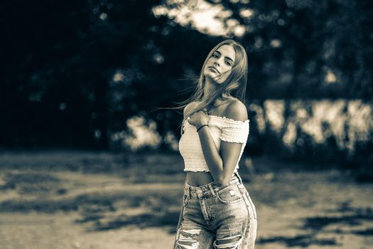 Beautiful teen girl wearing white bare shoulders top and jeans posing against trees on blurred nature background at river side. Black and white tinted image.