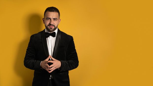 Handsome man in tuxedo standing fingers folded down smiling on camera, work profession lifestyle. Handsome young smiling caucasian man isolated on yellow background. 