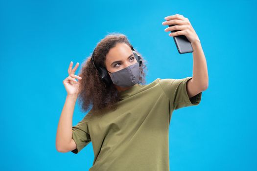 Dancing African American girl listen music on her smartphone with headphones in olive t-shirt, reusable face mask, to prevent others from corona COVID-19 infection isolated on blue background. 