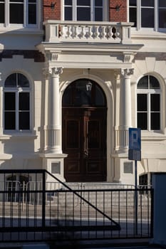 View of the front wall of the building with the main entrance. Unadorned canopy door with a richly decorated balcony and two columns.