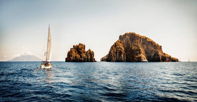 Modern white sailboat sailing slowly in rippling sea near rough rock in cloudless day on resort in Italy, Eolie islands