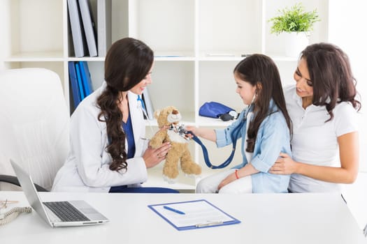Cute small child patient pretending to be doctor, holding teddy bear toy at meeting with general practitioner. Friendly nurse showing how stethoscope works to smiling little girl at checkup in clinic.