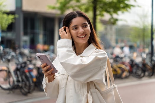 Sustainable young multiethnic woman wearing white eco clothes and a shopper holding a phone walking in the street filled with bicycles in the background.